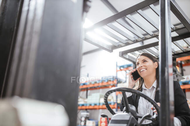 Cheerful female manager talking on mobile phone while operating forklift at warehouse — Stock Photo