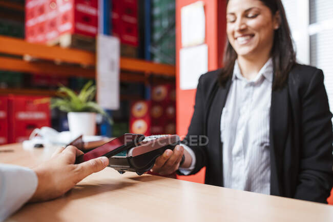 Gerente sonriente sosteniendo lector de tarjetas para el cliente con teléfono inteligente - foto de stock