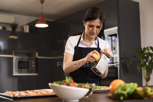 Woman cooking in kitchen pouring grated lemon on a dish — Stock Photo