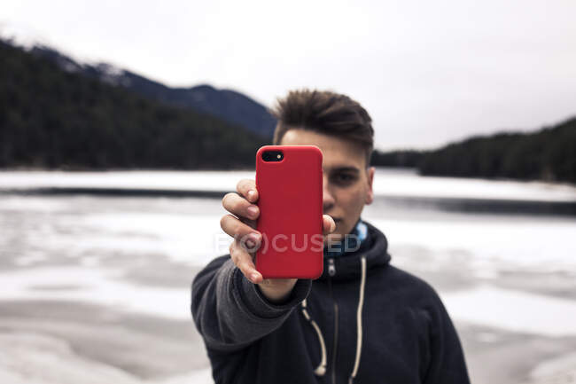 Young man holding red cell phone at a lake in winter — Stock Photo