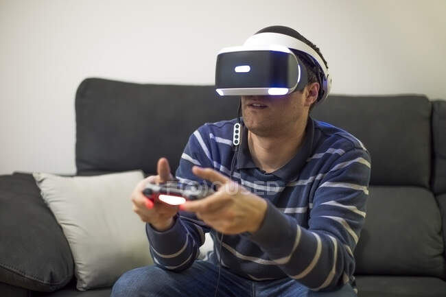 Joven con gafas VR sentado en el sofá en casa jugando videojuego - foto de stock