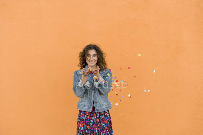 Portrait of smiling young woman throwing confetti in the air in front of orange wall — Stock Photo
