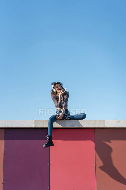 Giovane donna seduta su un muro ad ascoltare musica con le cuffie sul suo smartphone — Foto stock