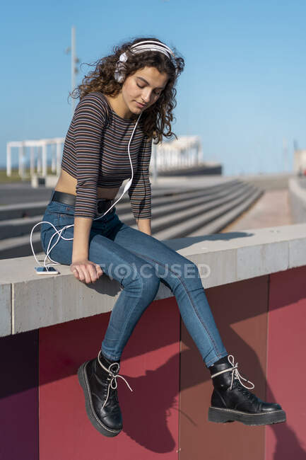 Young woman sitting on a wall listening music with headphones on her smartphone — Stock Photo