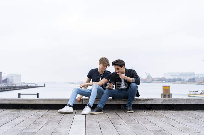 Dinamarca Copenhague dos jóvenes sentados en el paseo marítimo con teléfono celular - foto de stock