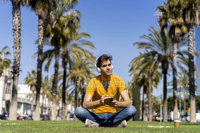 España, Barcelona, hombre sentado en el césped de la ciudad con auriculares y portátil - foto de stock