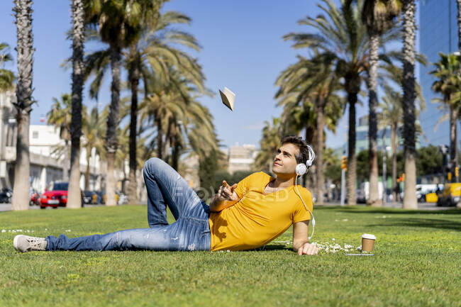 España, Barcelona, hombre tumbado en el césped de la ciudad con auriculares vomitando portátil - foto de stock