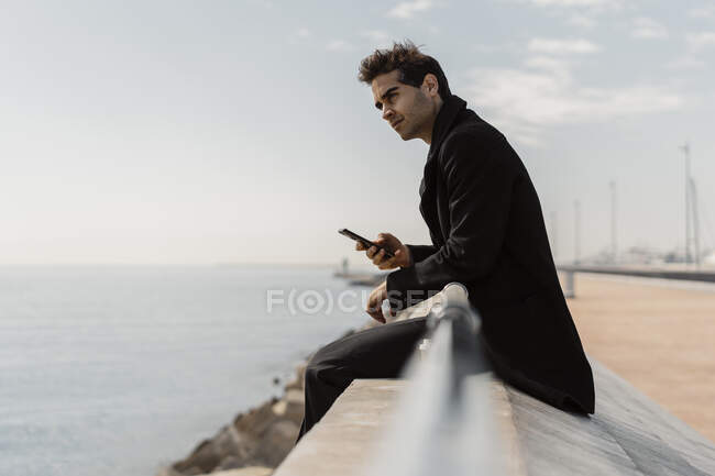 Uomo d'affari seduto sul muro di banchina con il telefono cellulare — Foto stock