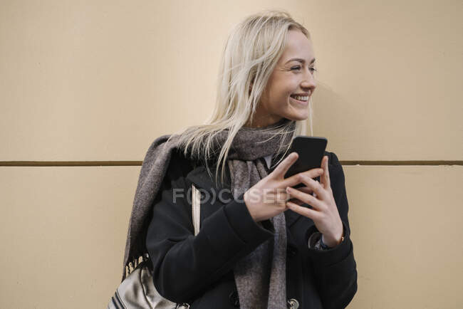Улыбающаяся молодая женщина с мобильным телефоном у стены — стоковое фото