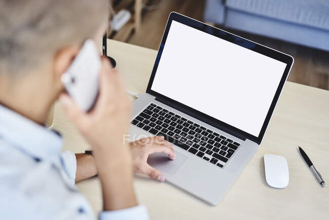 Young man talking on phone while typing on laptop in home office — Stock Photo