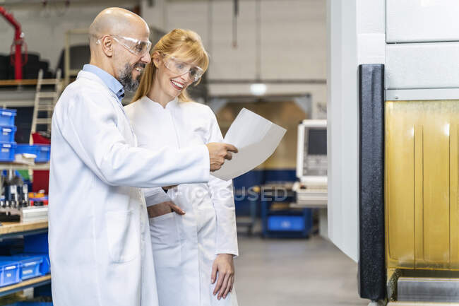 Two happy technicians wearing lab coats and safety glasses looking at plan in factory — Stock Photo