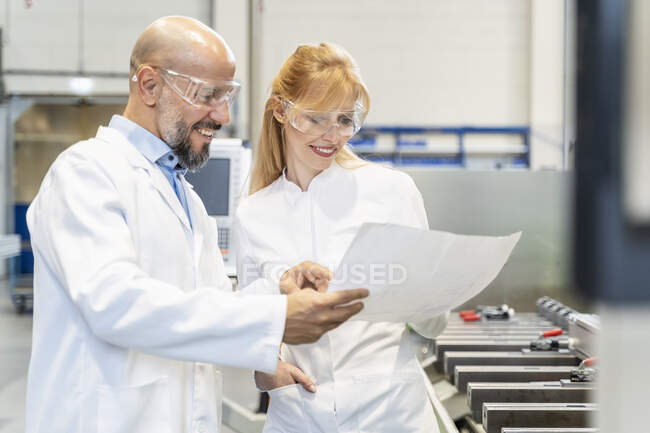 Zwei glückliche Techniker mit Laborkittel und Schutzbrille schauen sich den Plan in der Fabrik an — Stockfoto