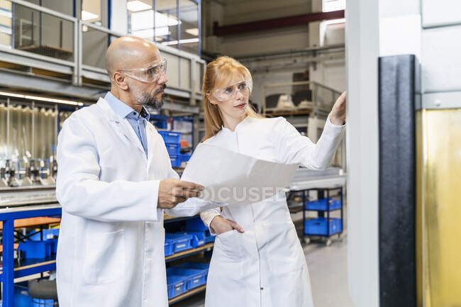 Two technicians wearing lab coats and safety glasses examining machine — Stock Photo