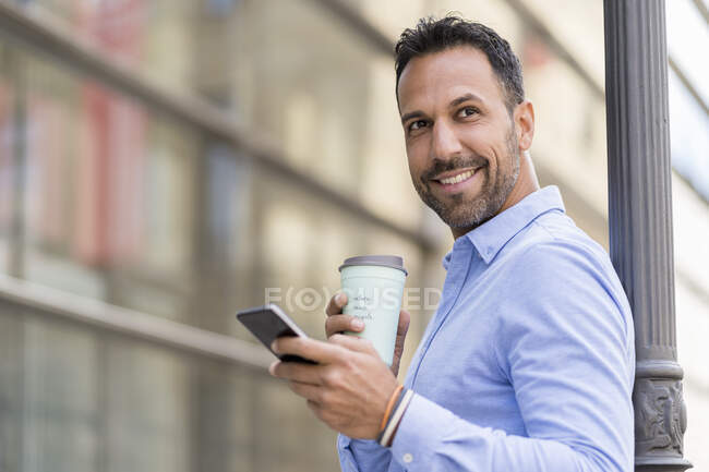 Улыбающийся бизнесмен с кофе на вынос и сотовым телефоном в городе — стоковое фото