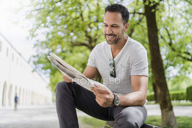Lächelnder Mann sitzt auf Parkbank und liest Zeitung — Stockfoto