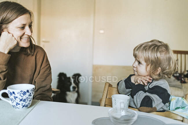 Smiling mother and daughter sitting at table at home with dog in background — Stock Photo
