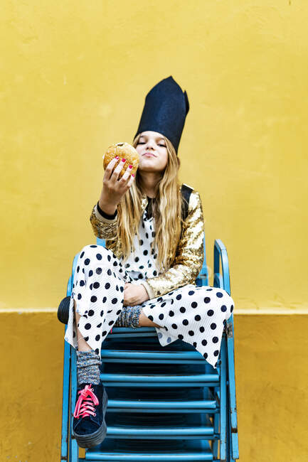 Girl wearing black crown sitting on stack of chairs holding hamburger — Stock Photo