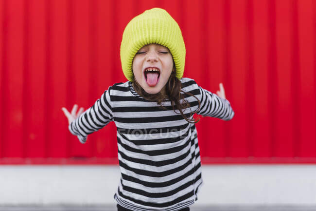 Portrait of little girl wearing striped shirt and yellow cap sticking out tongue in front of red background — Stock Photo