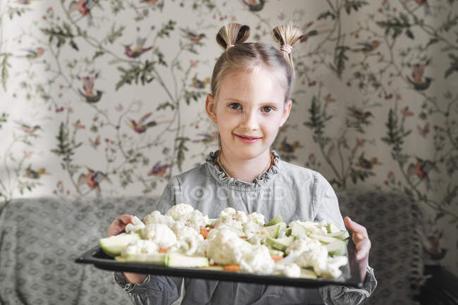 Portrait of blond girl with baking tray of raw vegetables — Stock Photo