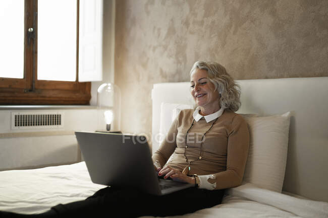 Ritratto di donna d'affari matura seduta sul letto a lavorare su laptop — Foto stock