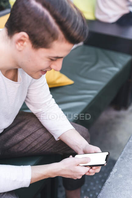 Smiling young man sitting on couch in a cafe using cell phone — Stock Photo