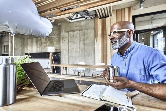 Mature businessman sitting at table in modern office with cloud balloon and laptop — Stock Photo