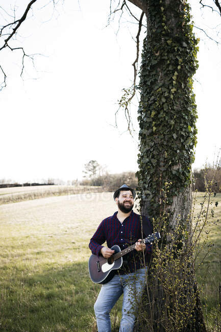 Hombre tocando la guitarra en un árbol en un prado - foto de stock