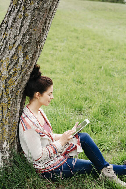 Junge Frau sitzt mit Tablet auf dem Land an einem Baum — Stockfoto