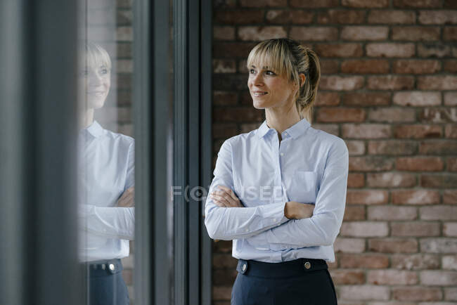 Femme d'affaires confiante regardant par la fenêtre, les bras croisés — Photo de stock