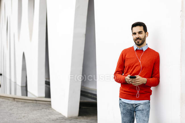 Homme d'affaires décontracté appuyé sur le mur, à l'aide d'écouteurs et smartphone — Photo de stock