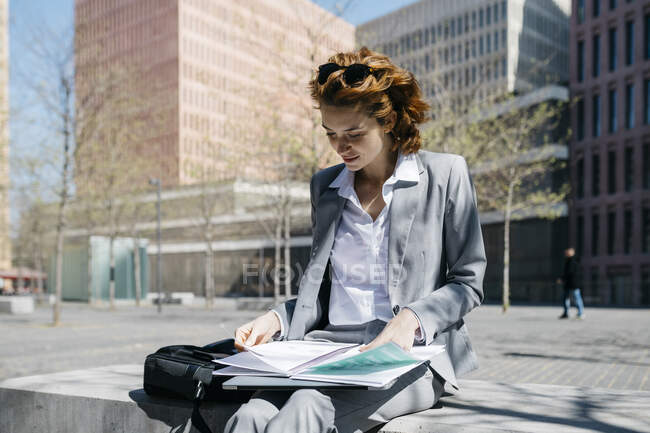 Joven empresaria leyendo documentos, sentada en un banco de la ciudad en un día soleado - foto de stock