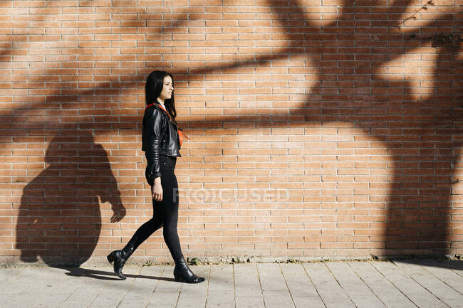 Молода жінка з червоною сумкою, що йде перед цегляною стіною і тінь дерева — стокове фото