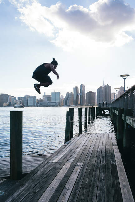 USA, New York, Brooklyn, young man doing Parkour jump from wooden pole in front of Manhattan skyline — Stock Photo