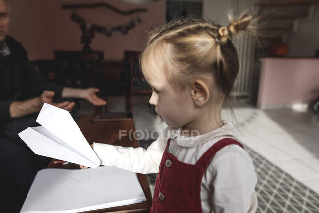 Girl holding paper plane in living room with grandfather in background — Stock Photo