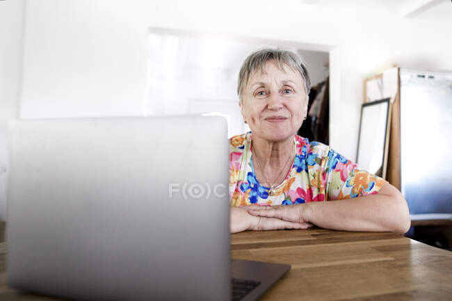 Portrait of smiling senior woman sitting at table at home using laptop — Stock Photo