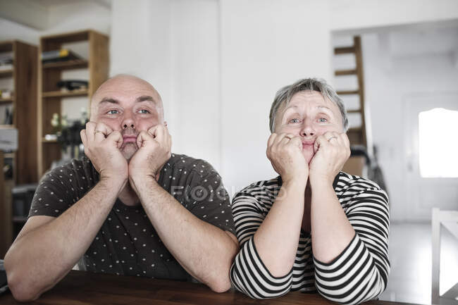Retrato de hijo adulto con madre mayor sentada a la mesa en casa con la cabeza en las manos - foto de stock