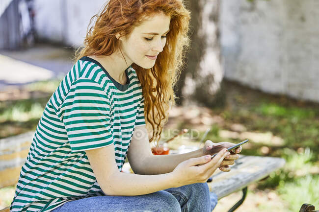 Smiling redheaded young woman  sitting on bench in the garden looking at cell phone — Stock Photo
