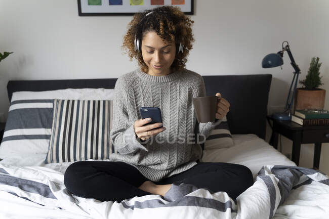 Жінка сидить на ліжку, п'є каву, слухає музику навушниками та смартфонами. — стокове фото