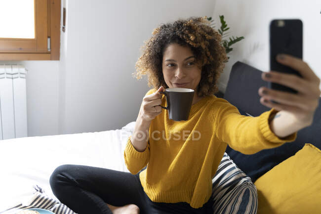 Жінка сидить на ліжку, бере селфі, п'є каву. — стокове фото