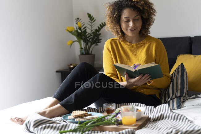 Mujer sentada en la cama, desayunando sano, leyendo un libro - foto de stock