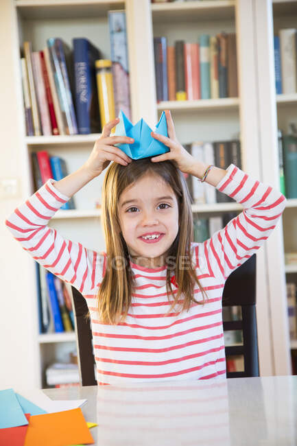 Retrato de una chica sonriente poniéndose un sombrero de papel hecho a sí misma en casa - foto de stock