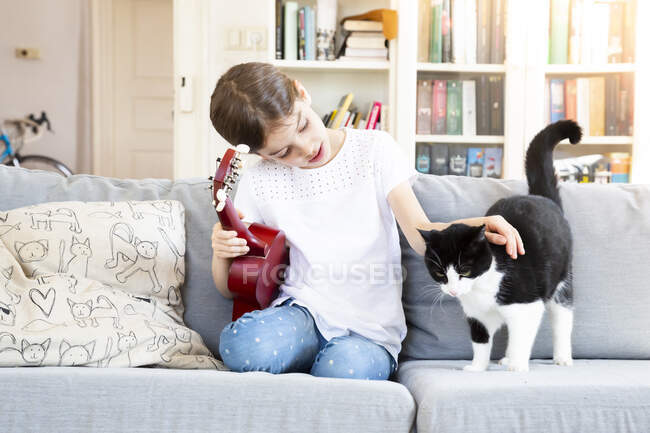 Chica con ukelele sentado en sofá acariciando gato - foto de stock