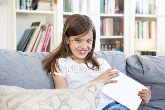 Porträt eines glücklichen kleinen Mädchens, das mit dem digitalen Tablet zu Hause auf der Couch sitzt — Stockfoto