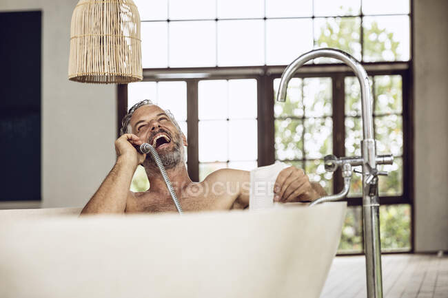 Riéndose hombre en bañera usando cabezal de ducha como receptor de teléfono - foto de stock
