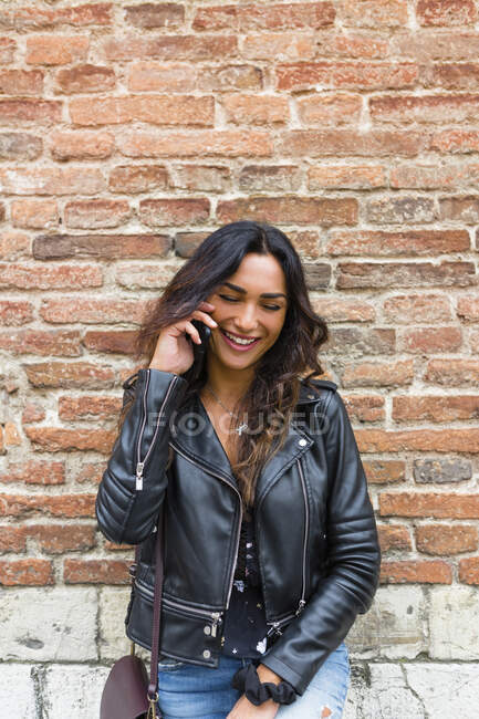 Retrato de mulher jovem vestindo jaqueta de couro preto, usando smartphone, parede de tijolo no fundo — Fotografia de Stock