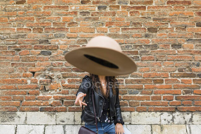Retrato de jovem mulher vestindo jaqueta de couro preto, parede de tijolo, jogando chapéu — Fotografia de Stock