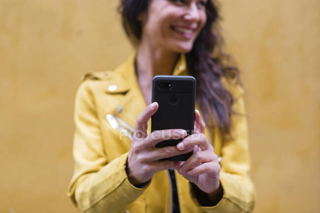 Retrato de jovem mulher vestindo jaqueta de couro amarelo e tirando uma selfie, parede amarela no fundo — Fotografia de Stock