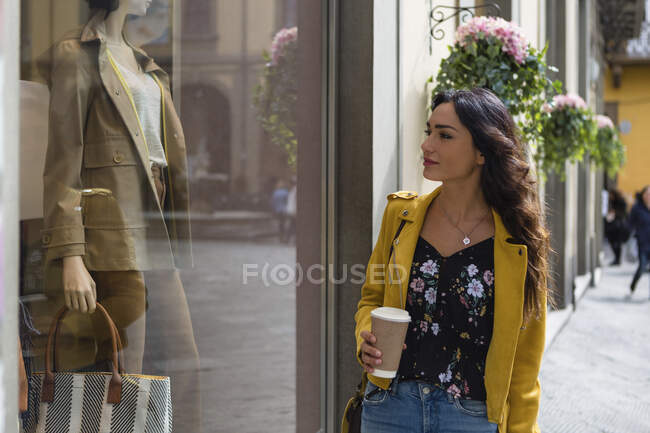 Retrato de jovem mulher vestindo jaqueta de couro amarelo, segurando xícara de café durante as compras — Fotografia de Stock