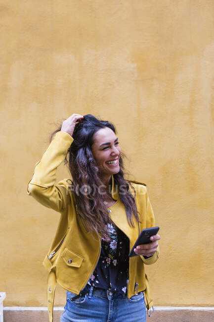 Retrato de jovem mulher rindo usando jaqueta de couro amarelo e segurando smartphone — Fotografia de Stock