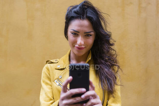 Retrato de jovem mulher vestindo jaqueta de couro amarelo e tirando uma selfie, parede amarela no fundo — Fotografia de Stock